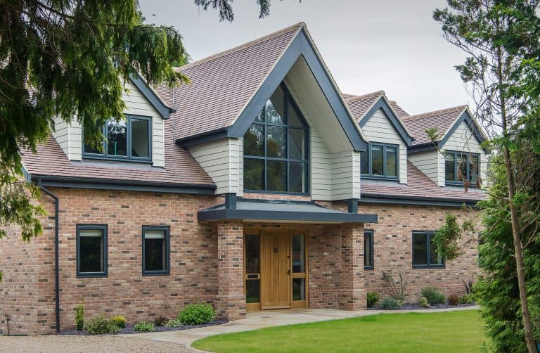 Modern two-story brick house with large windows, a wooden front door, and a peaked roof. The house is surrounded by green lawn, shrubs, and trees.