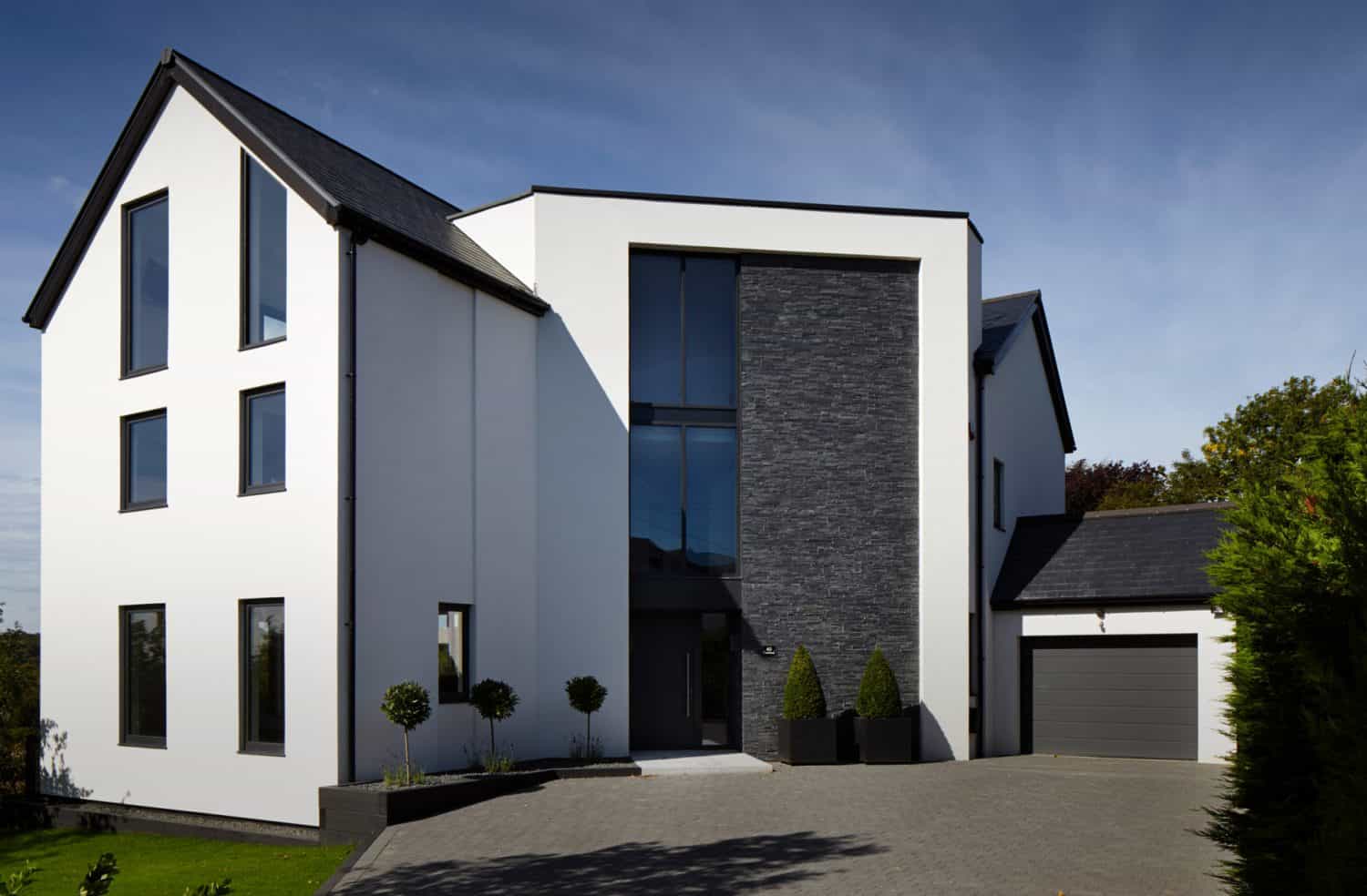 Modern two-story house with large windows, white exterior walls, dark stone accents, and a gray garage. The driveway is paved and landscaped with small trees and shrubs. The sky is clear and blue.