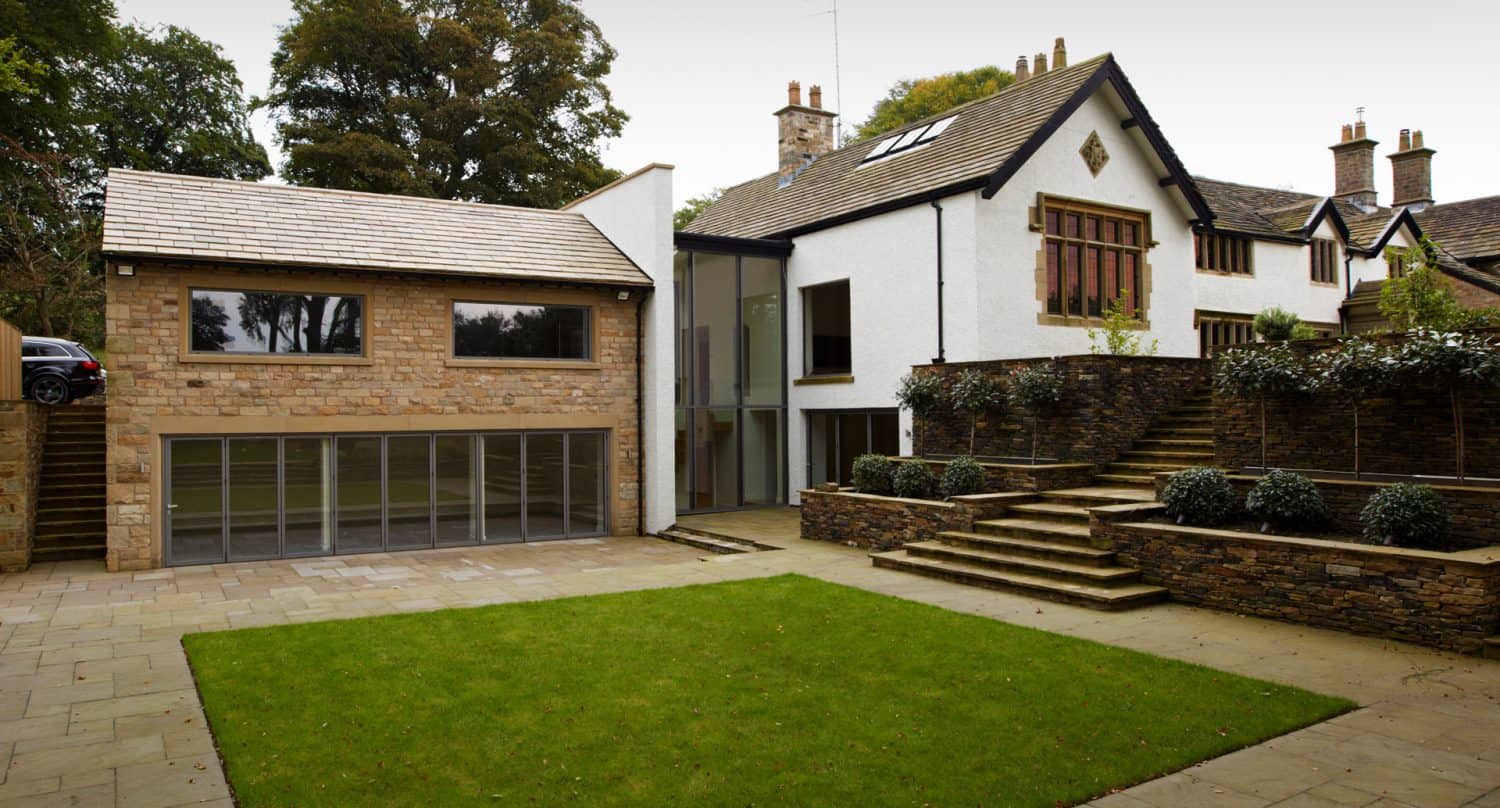 Modern extension with glass doors is attached to a traditional white house with stone accents. There are stone steps, a retaining wall with plants, and a small rectangular lawn in the courtyard.