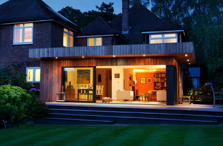 A modern house with large glass doors opens to a warmly lit living and dining area at dusk. The garden lawn and lush greenery are visible in the foreground.