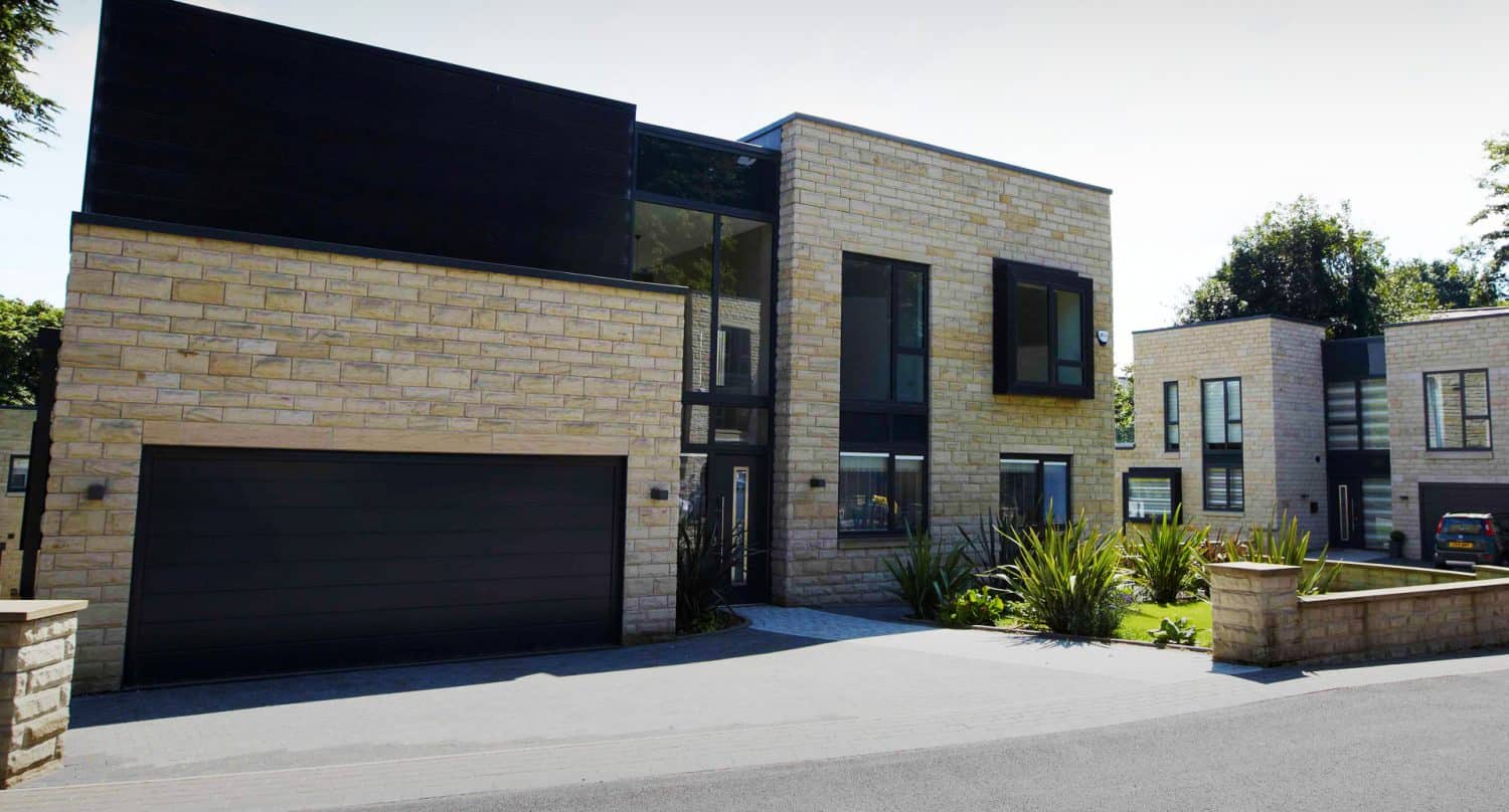 Modern two-story house with light stone exterior, large black-framed windows, and a double garage. The driveway is paved, with landscaped plants in front. Another similar building and a car are visible in the background.