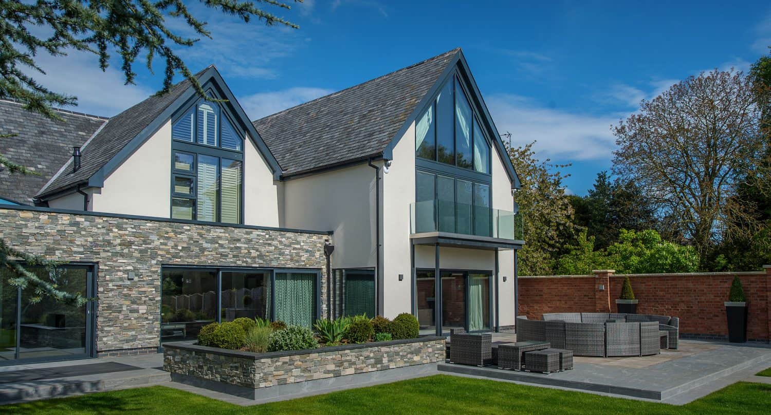Modern two-story house with large windows, stone and white exterior, sloped roof, balcony, and a landscaped patio with outdoor seating, surrounded by greenery and a brick wall under a blue sky.