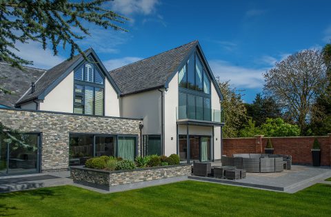 Modern two-story house with large windows, stone and white exterior, sloped roof, balcony, and a landscaped patio with outdoor seating, surrounded by greenery and a brick wall under a blue sky.