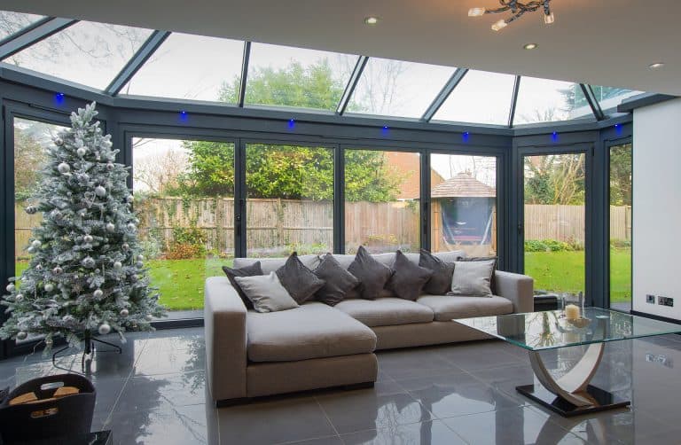 A modern sunroom with large glass windows, a light grey sectional sofa with many pillows, a glass coffee table, and a decorated Christmas tree. Outside, a green garden and wooden fence are visible.