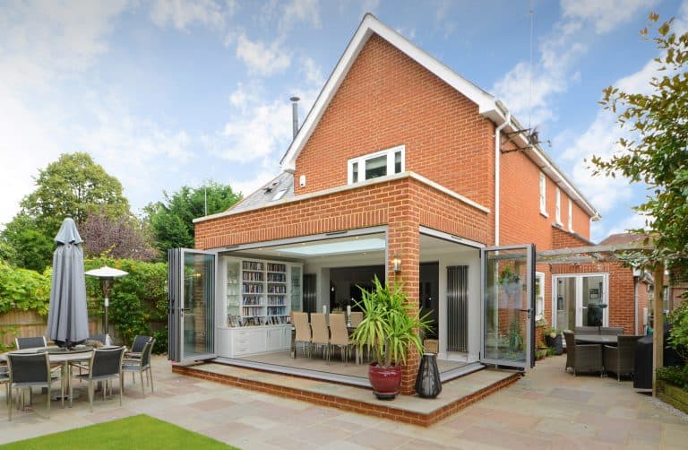 Modern brick house with large glass folding doors opening to a patio, revealing a dining area and bookshelf inside. Outdoor seating areas and green potted plants surround the patio. Sky is blue with scattered clouds.