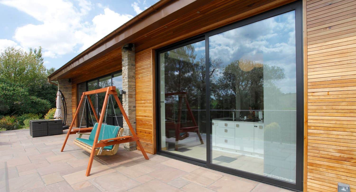 A modern house with wooden siding and large sliding glass doors, reflecting trees and sky. A wooden patio swing with cushions sits on a stone patio next to the house.
