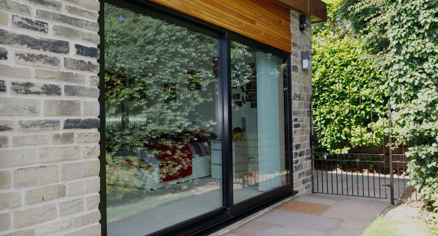A large glass sliding door reflects trees and greenery outside. Through the glass, a cozy bedroom with a bed and cushions is visible. The house has stone walls and a small patio with paving stones.