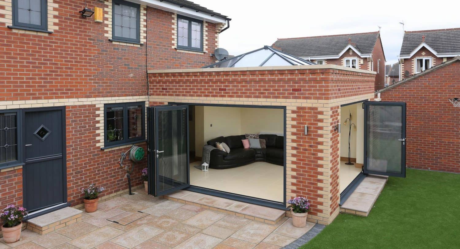 A modern brick house with large glass folding doors opening to a living area with a corner sofa. The patio is tiled, and there is a small grass lawn with potted plants by the entrance.