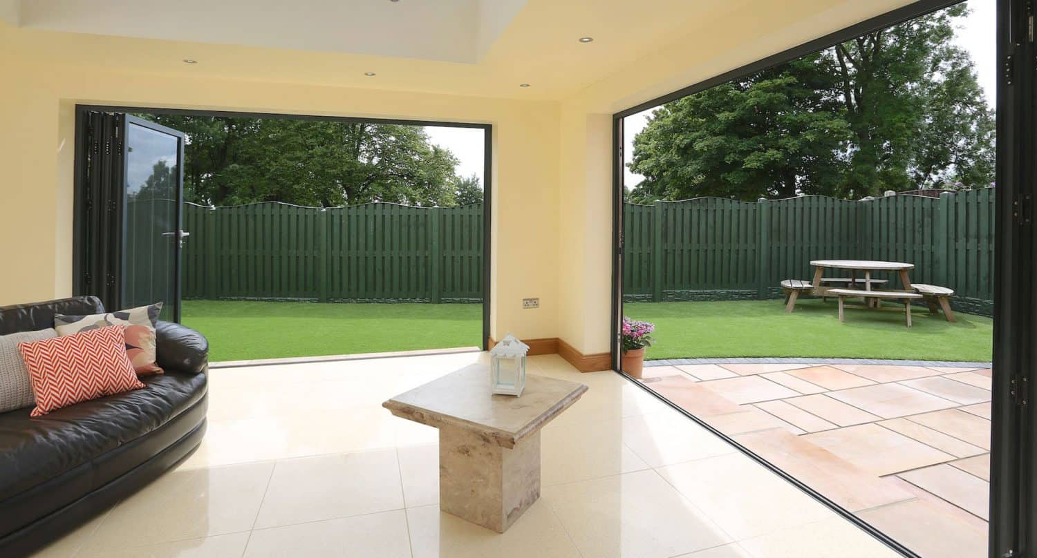 Modern sunroom with open folding glass doors leading to a garden. Inside, theres a black sofa with cushions and a small marble table. Outside, there’s artificial grass, a green fence, and a wooden picnic bench.