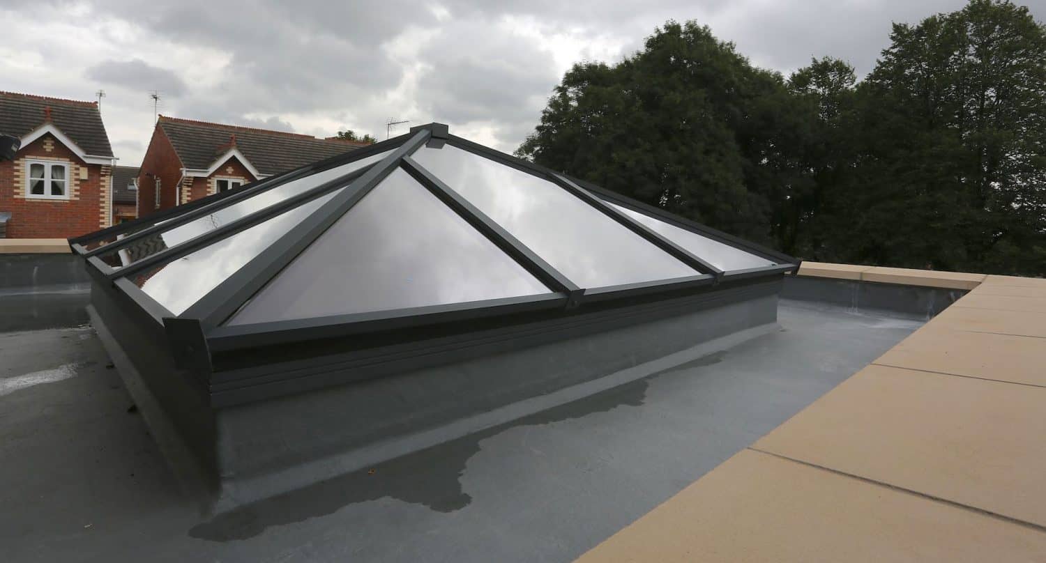 A modern glass pyramid-shaped skylight is set into a flat rooftop, reflecting the cloudy sky above. Nearby are trees and the upper floors of brick houses with white-framed windows.