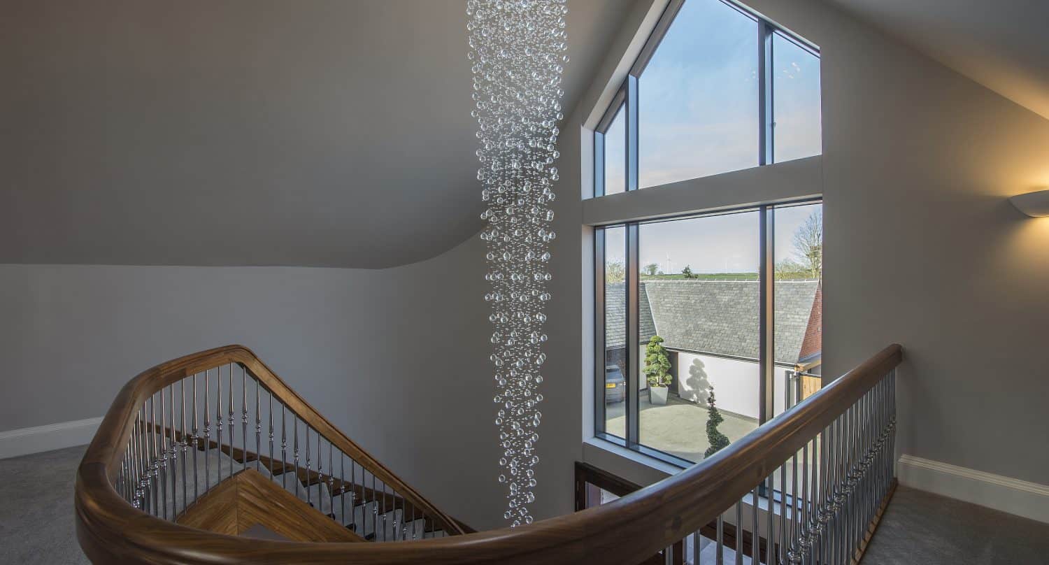 A modern staircase with a wooden handrail and metal balusters curves around a tall, glass crystal chandelier. Large triangular windows and bespoke glass solutions let in natural light, revealing rooftops and trees outside.