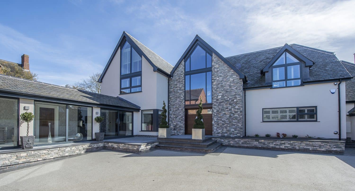 Modern two-story house with large glass windows, gray stone accents, and a spacious paved driveway on a clear, sunny day. The entrance features potted plants, geometric architecture, and sleek sliding doors that enhance the airy design.