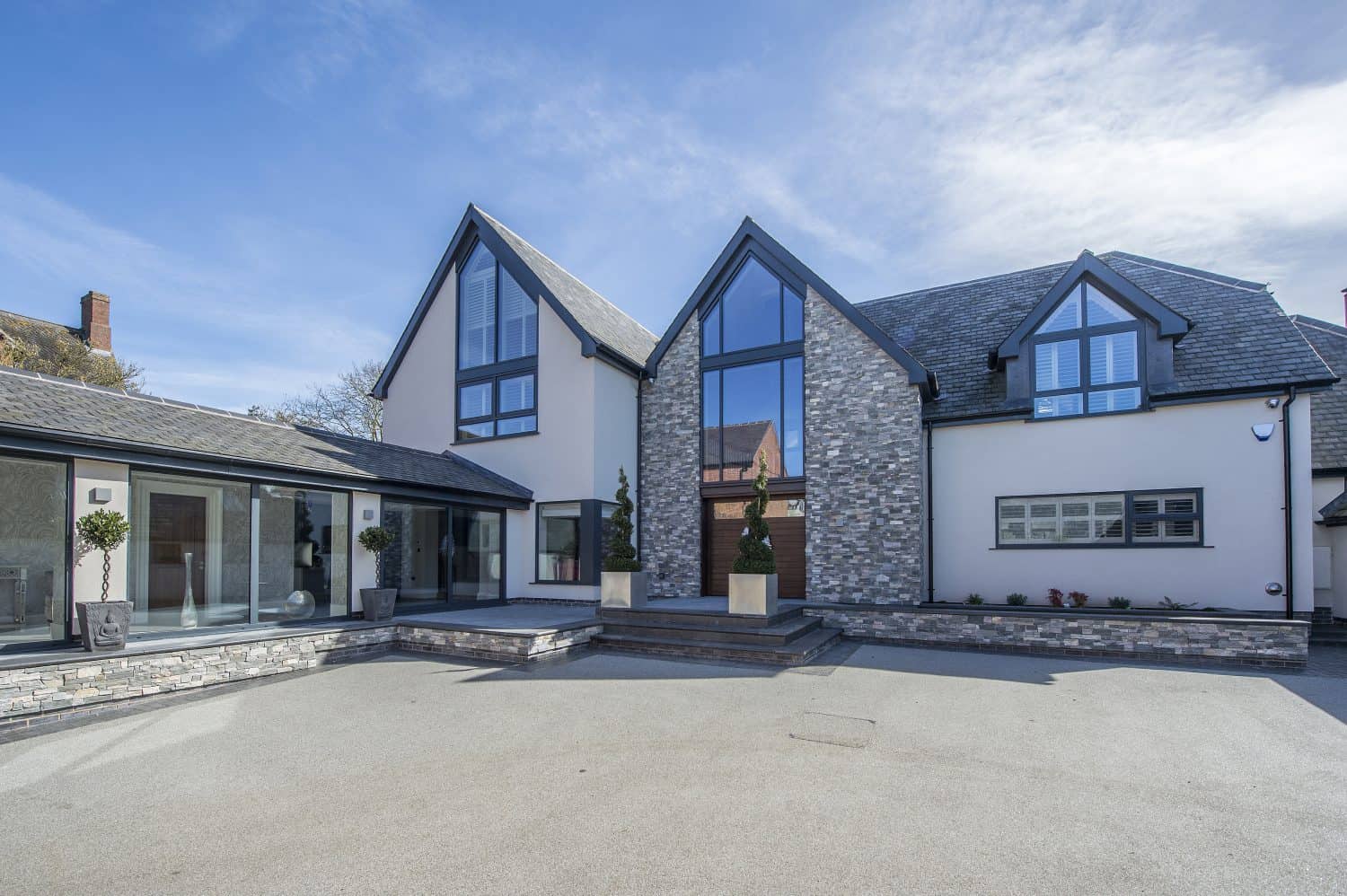 Modern two-story house with large glass windows, gray stone accents, and a spacious paved driveway on a clear, sunny day. The entrance features potted plants, geometric architecture, and sleek sliding doors that enhance the airy design.
