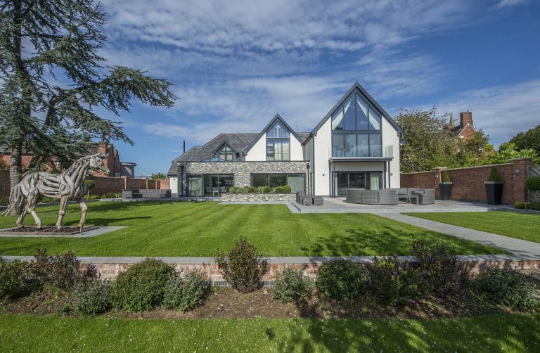 Modern two-story house with large glass windows, sliding doors, and triangular roof lines, set behind a manicured lawn and garden. A metal horse sculpture stands on the left side; the sky is bright with scattered clouds.