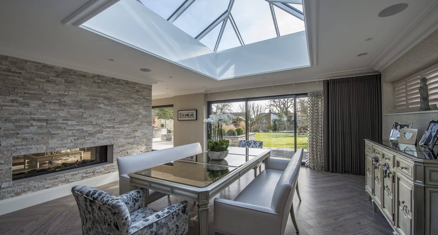 Modern dining room with a glass ceiling, mirrored table, white benches, patterned armchair, stone fireplace, large windows with bespoke glass solutions, and a sideboard displaying decor, overlooking a green garden.