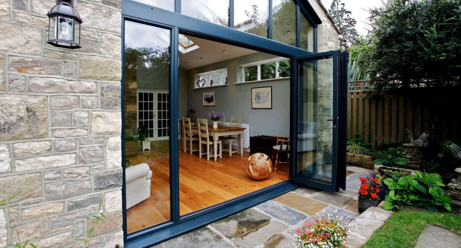A modern dining area with wooden floors and a table is visible through large, open glass doors in a stone-walled house. A garden with flowers and greenery surrounds the patio outside.