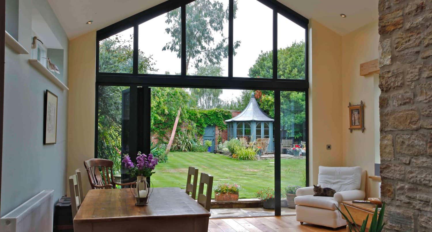 A dining area with wooden table and chairs sits beside a large floor-to-ceiling glass window overlooking a lush green garden with a gazebo. A white armchair and stone wall are on the right.