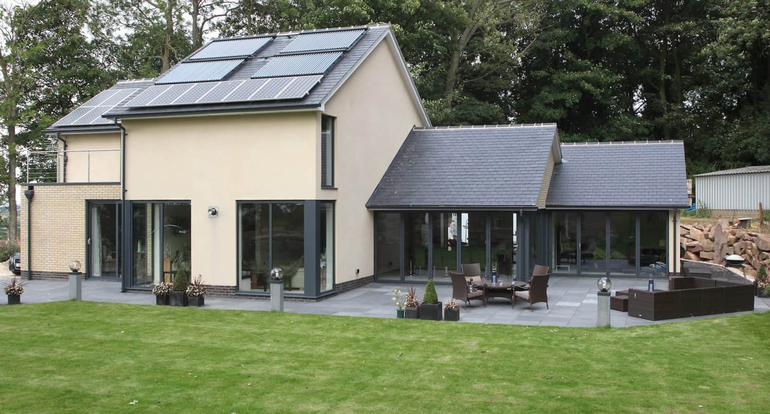 Modern two-story house with solar panels on the roof, large glass doors, and a spacious patio with outdoor furniture. The property is surrounded by a well-kept lawn and trees in the background.