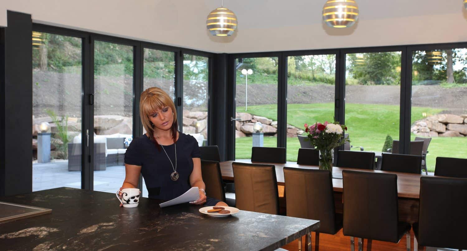 A woman holding a cup and reading a paper stands at a kitchen island with a plate of cookies. Behind her, a dining table and chairs face large windows overlooking a garden and patio area.