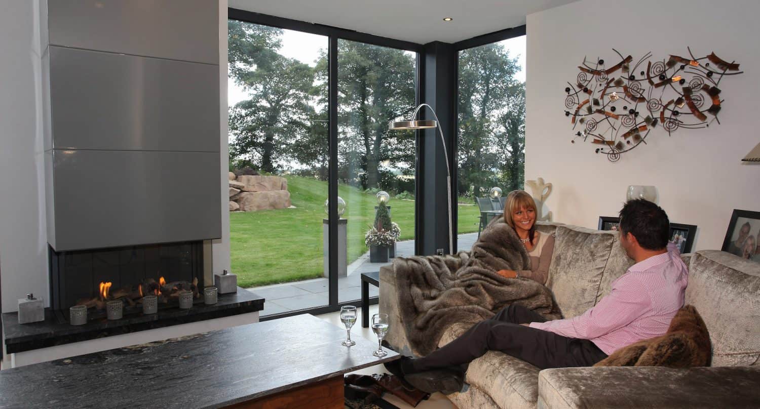 A couple sits on a cozy couch by a modern fireplace, wrapped in a blanket, smiling at each other. Large windows offer a view of a green lawn and trees outside. Two glasses of wine sit on the coffee table.