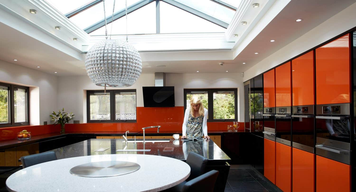 A modern kitchen with glossy orange cabinets, a large round island with a white countertop, a metallic spherical pendant light, and a woman standing by the window under a glass ceiling.