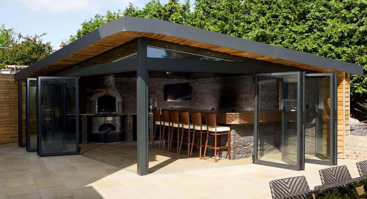 Modern outdoor kitchen with large glass doors, a bar area with high chairs, a built-in brick oven, and surrounding greenery. The structure has a sleek, angled roof and stone-textured walls.
