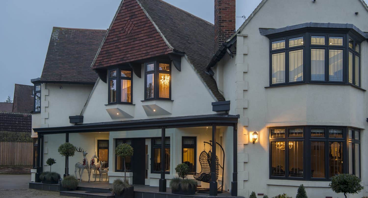 A large, modern two-story house with white walls, black trim, bay windows, and a covered porch featuring bespoke glass solutions and hanging chairs with potted plants, all warmly lit during dusk.