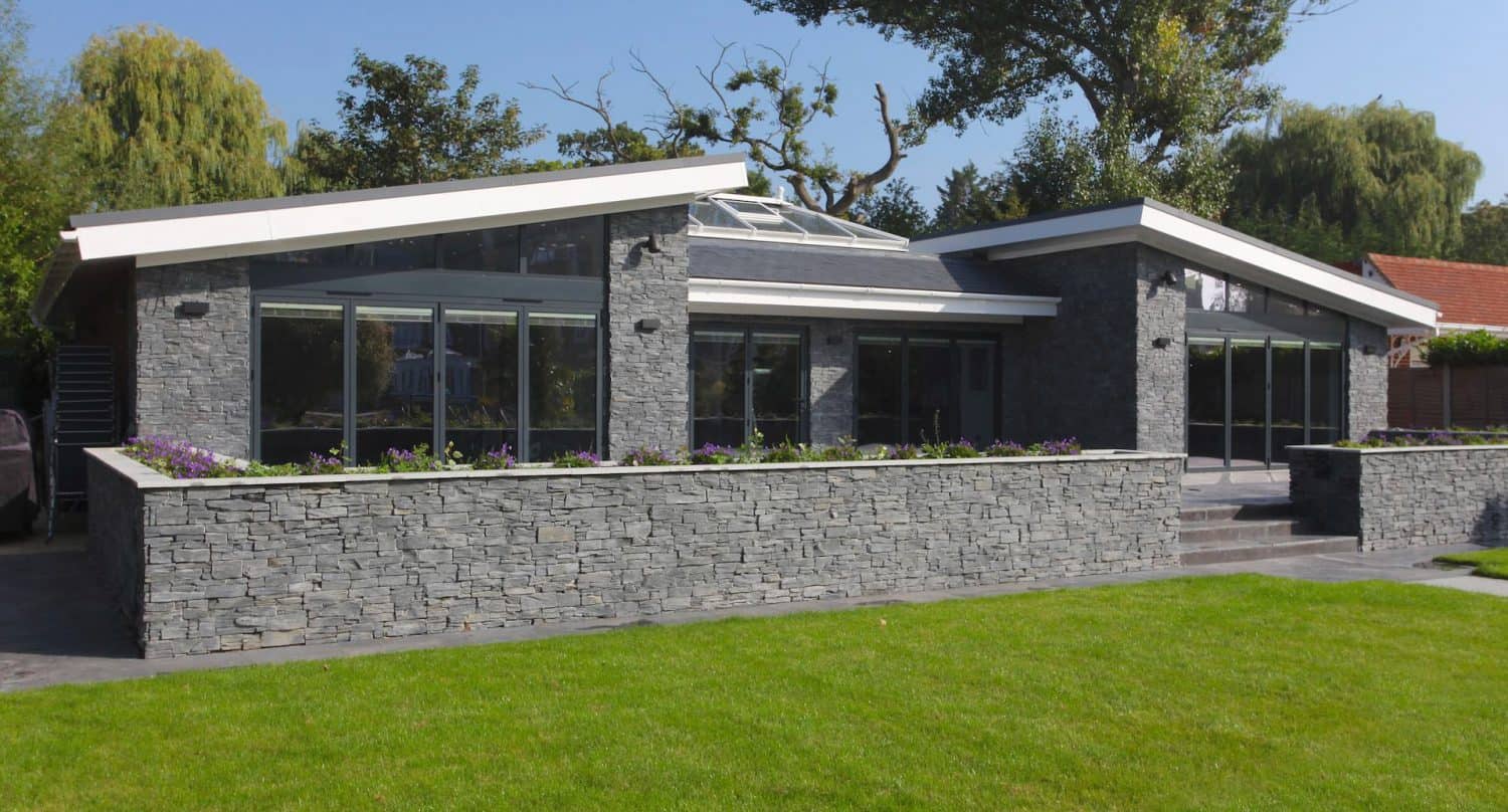 Modern house with large glass windows, stone exterior walls, sloped roofs, and a skylight. A well-maintained green lawn and a flower bed border the front of the building. Trees are visible in the background.