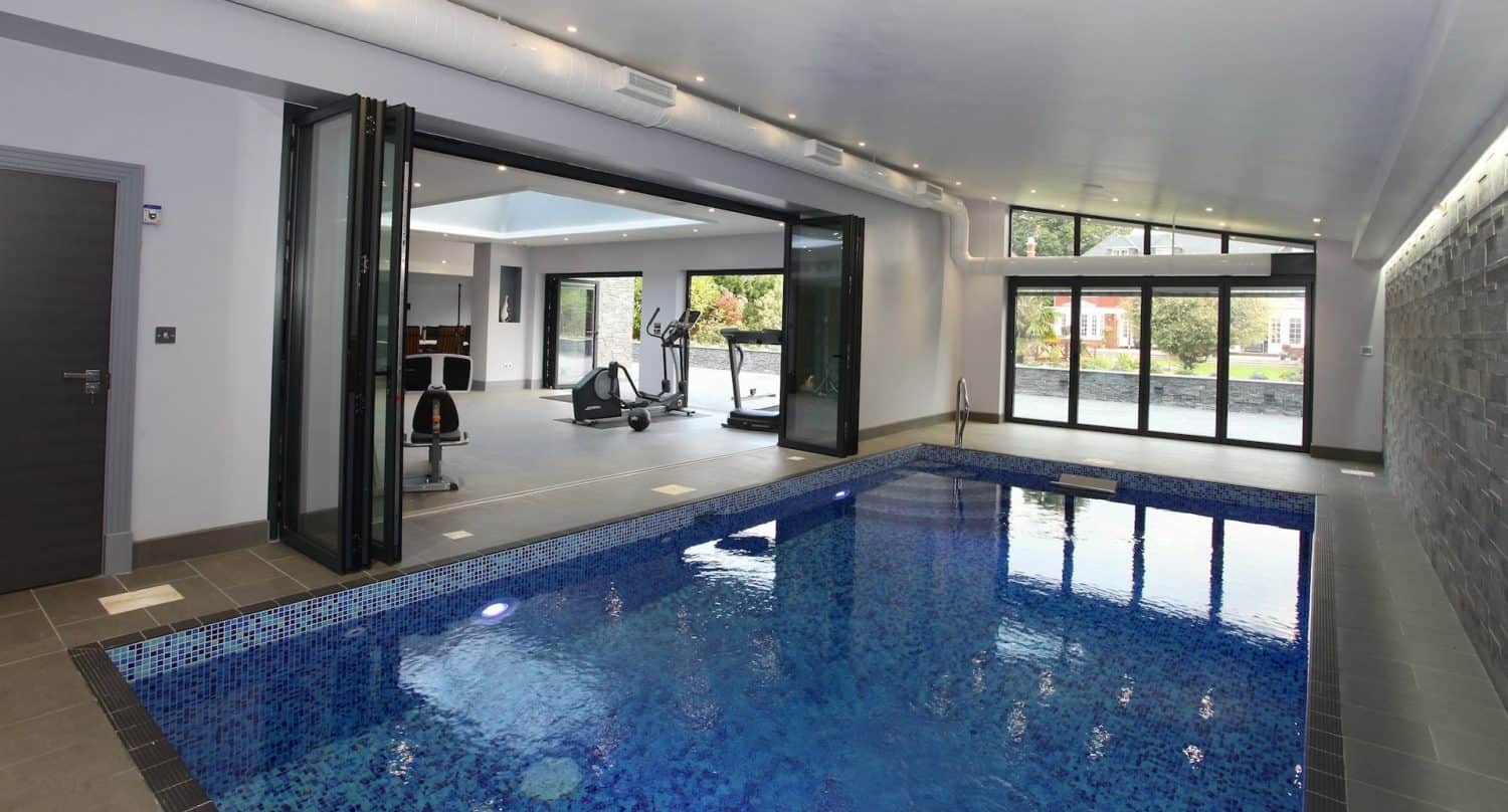 Indoor swimming pool with blue tile surround, beside a gym area with exercise machines. Large glass folding doors are open, connecting the pool space to the gym and allowing natural light from outside.