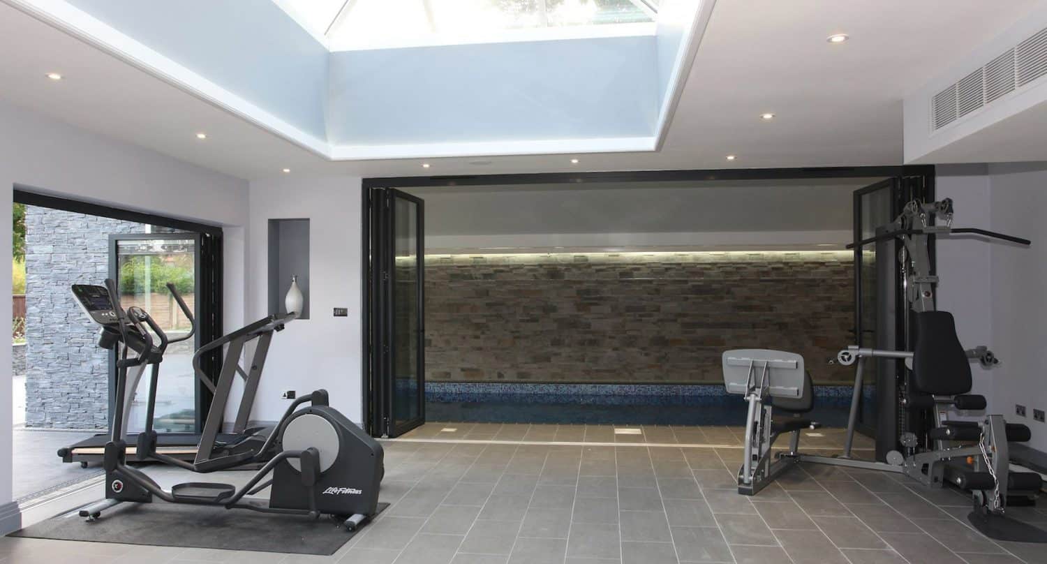 A modern home gym with cardio machines, strength equipment, large windows, and folding glass doors opening to an indoor swimming pool with stone accent wall. Natural light enters through a skylight.