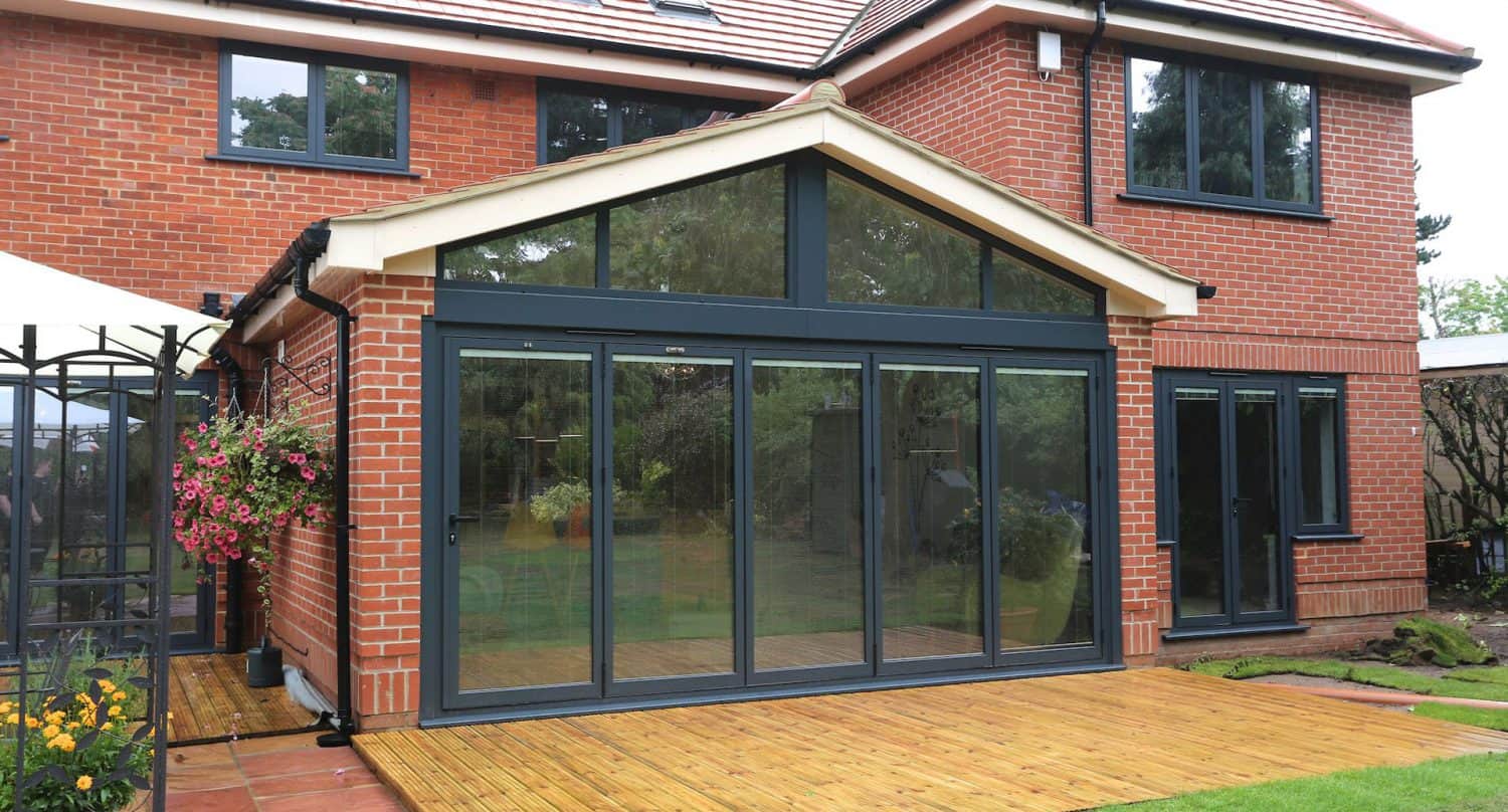 A modern red-brick house features a large glass extension with black-framed sliding doors opening onto a wooden deck, surrounded by a garden with flowers and greenery.