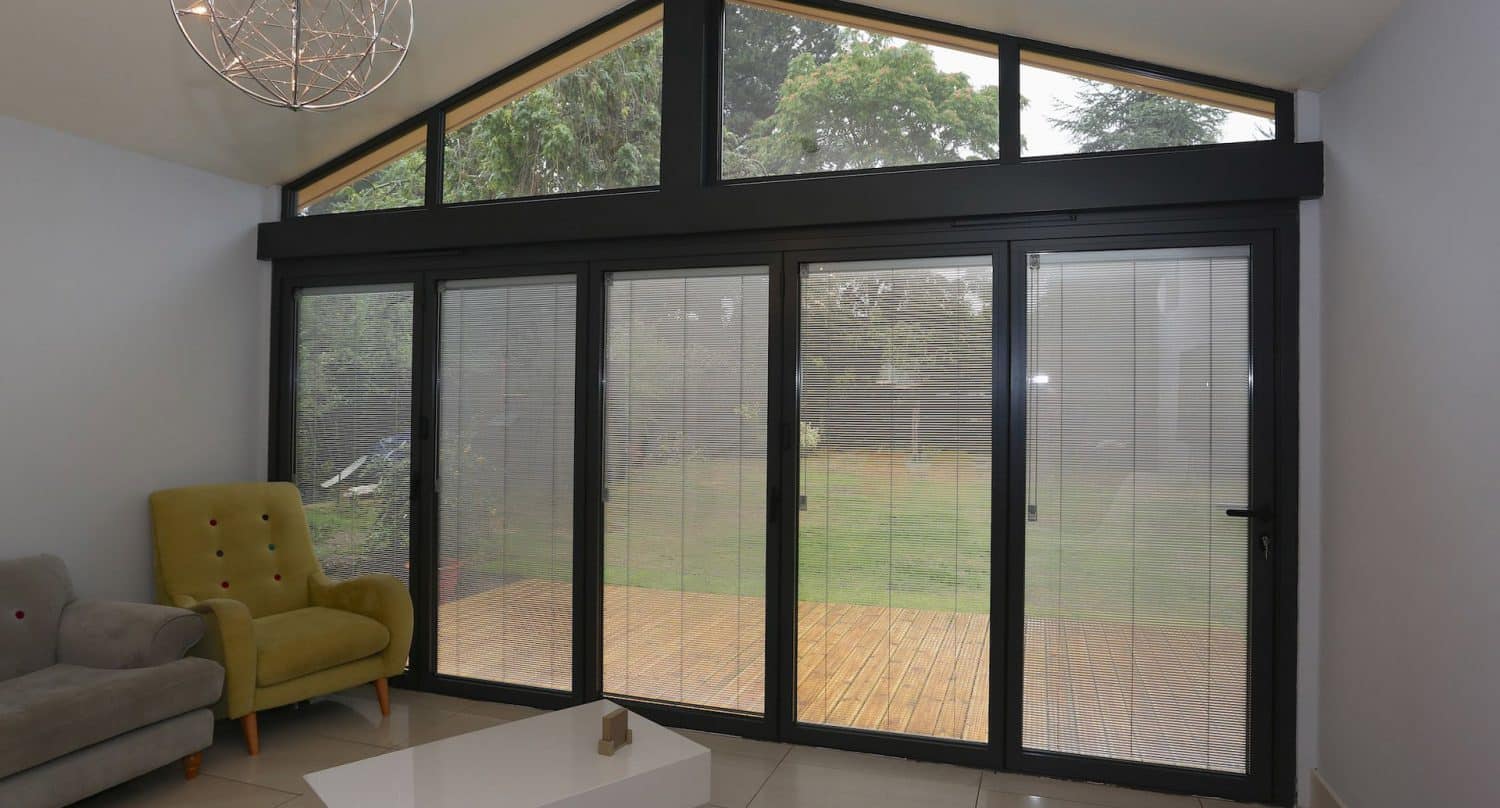 A modern living room with a yellow armchair, gray sofa, white coffee table, and large black-framed glass doors with blinds, opening to a wooden deck and green garden outside.
