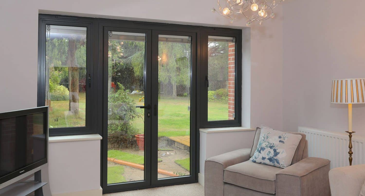A cozy living room with a gray armchair, a patterned lamp, a TV, and large glass patio doors offering a view of a green garden with trees outside.