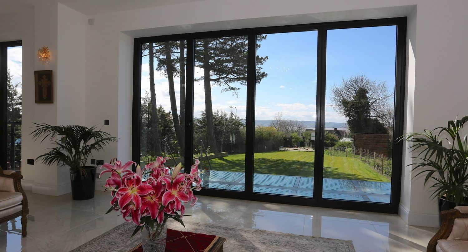 A bright living room with large sliding glass doors opens to a green backyard with trees. Indoors, there are potted plants, chairs, and a vase of pink lilies on a table. Blue sky and clouds are visible outside.