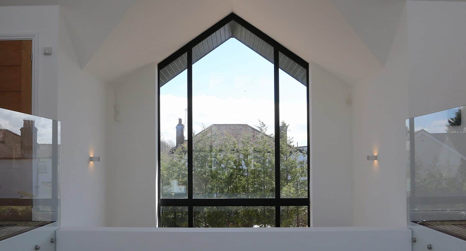 Large triangular window with black frame in a modern white room, offering a clear view of trees and rooftops outside under a blue sky with scattered clouds.