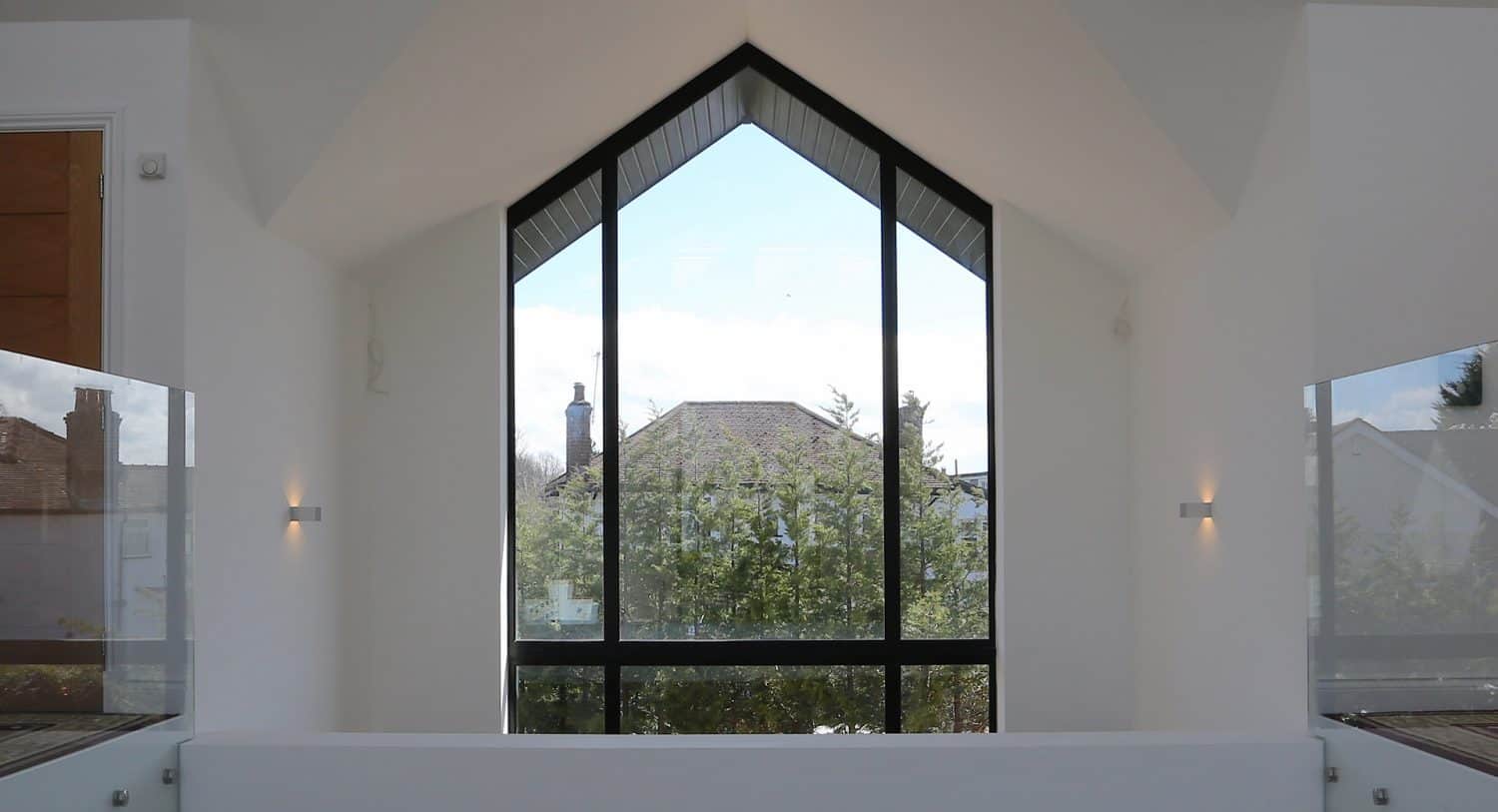 Large triangular window with black frame in a modern white room, offering a clear view of trees and rooftops outside under a blue sky with scattered clouds.