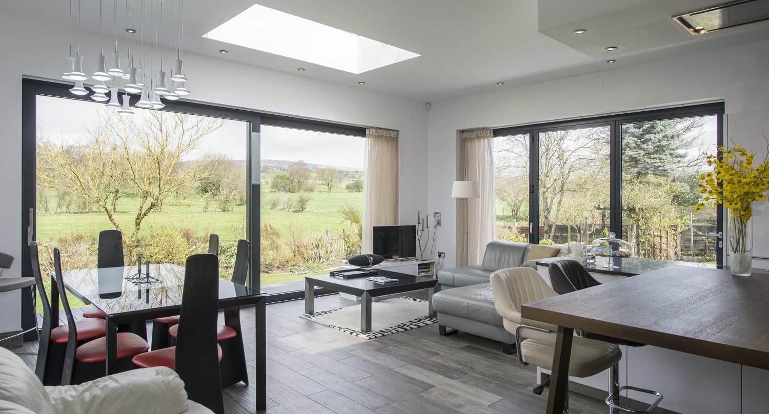 Modern living and dining area with large glass windows and bifold doors revealing a scenic rural landscape, featuring contemporary furniture, a skylight, wood flooring, and bright natural light.