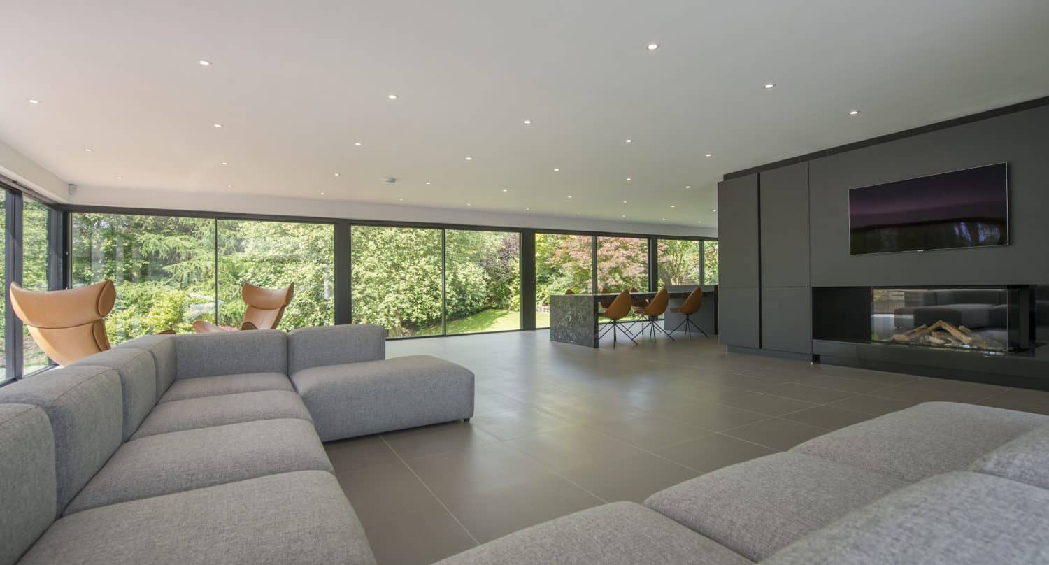 Spacious modern living room with a large gray sectional sofa, floor-to-ceiling sliding doors overlooking green trees, stylish chairs, a dark feature wall with a TV, and a sleek fireplace.