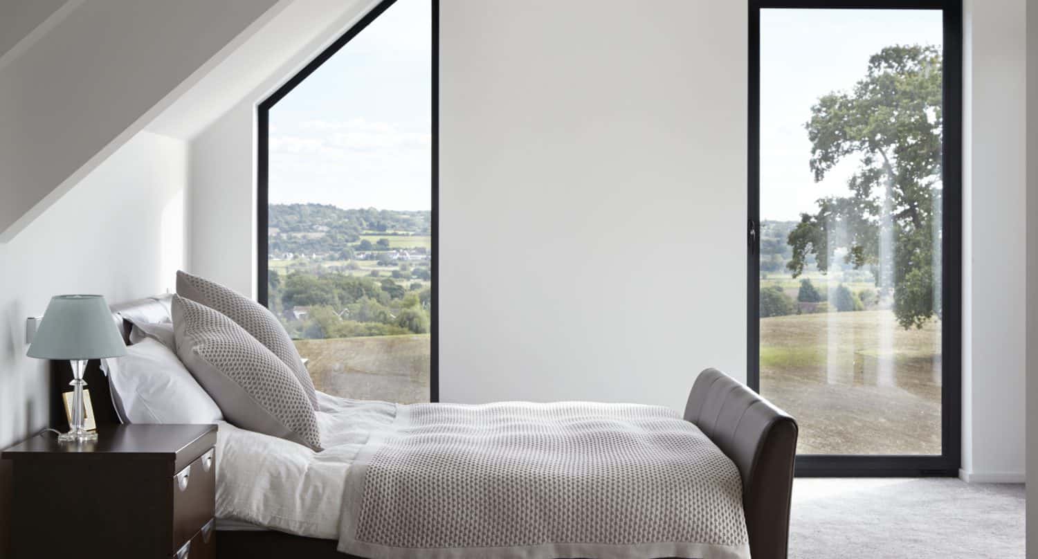 A modern, minimalist bedroom with a double bed, brown leather frame, beige bedding, nightstand with a lamp, and large windows featuring bespoke glass solutions that offer a scenic view of grassy fields and trees.