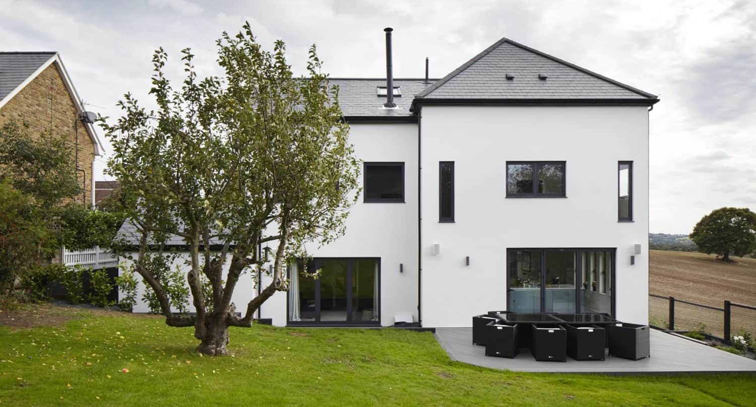 Modern white two-story house with large windows and bifold doors, a black outdoor dining set on a patio, a tree in the grassy backyard, and open fields in the background under a cloudy sky.