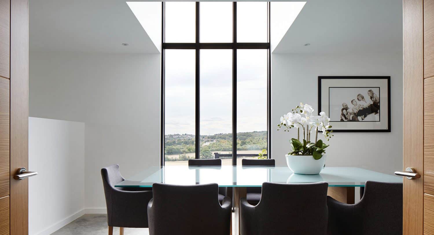 A modern dining room with a glass table, black chairs, a potted white orchid centerpiece, large floor-to-ceiling windows, and a framed black-and-white photo on a white wall. Natural light fills the room.