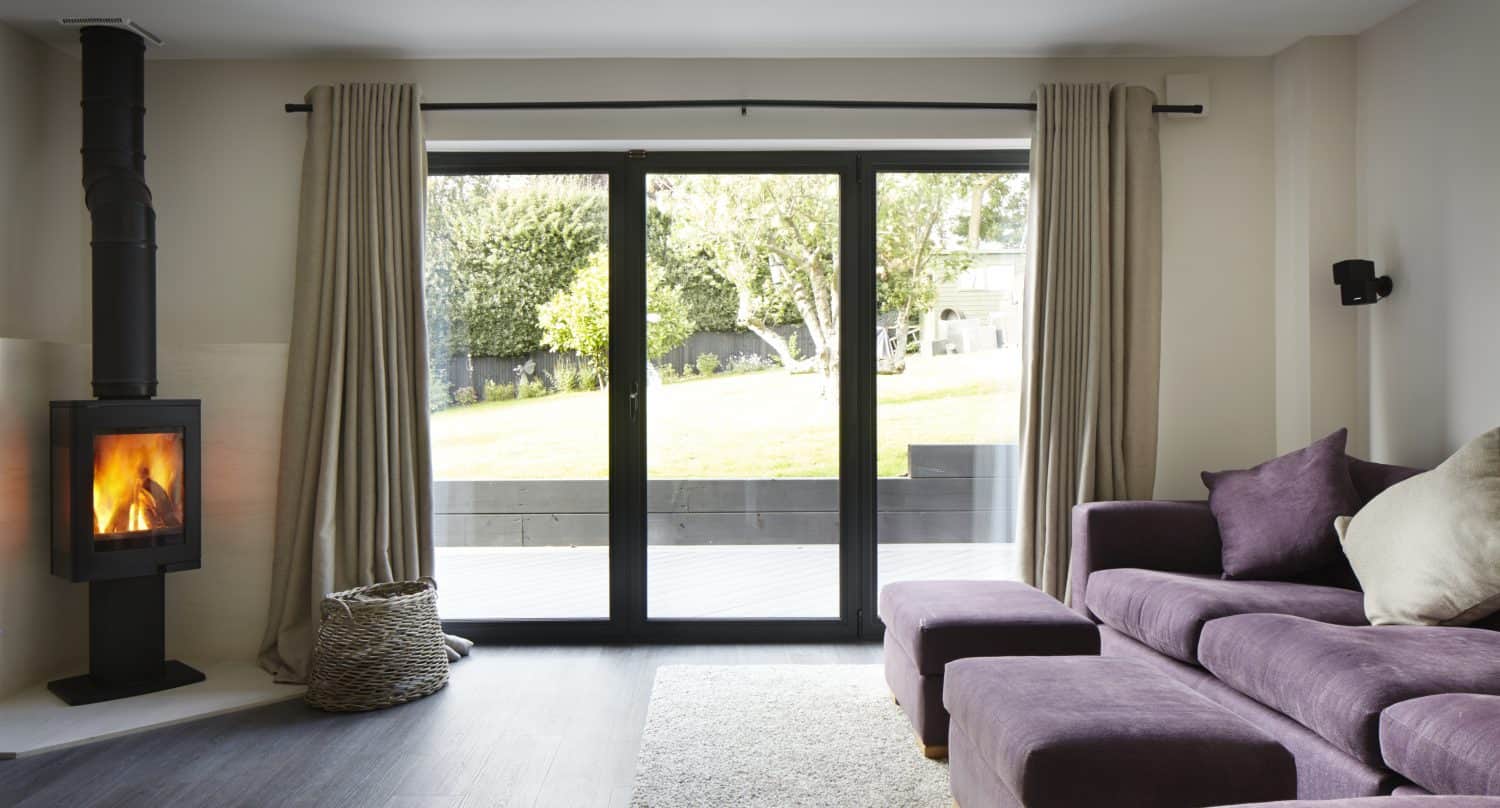 Modern living room with a purple sectional sofa, wood-burning stove, woven basket, light rug, and large bifold doors with beige curtains offering a beautiful view of the sunny garden outside.