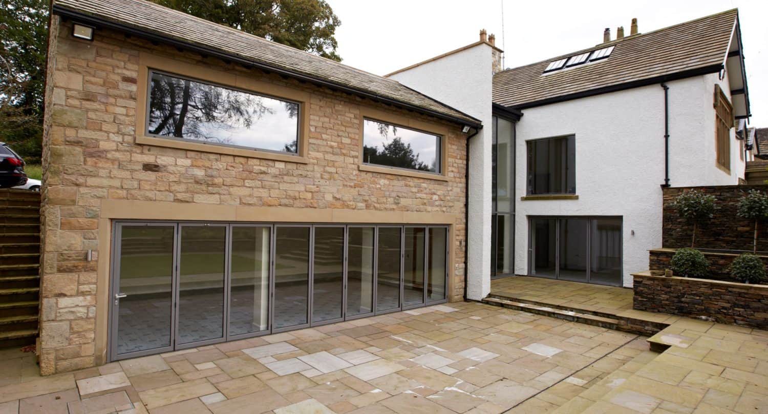 Modern house with stone and white exterior, large glass doors, and big windows facing a spacious paved patio; steps and greenery are visible on the left side.