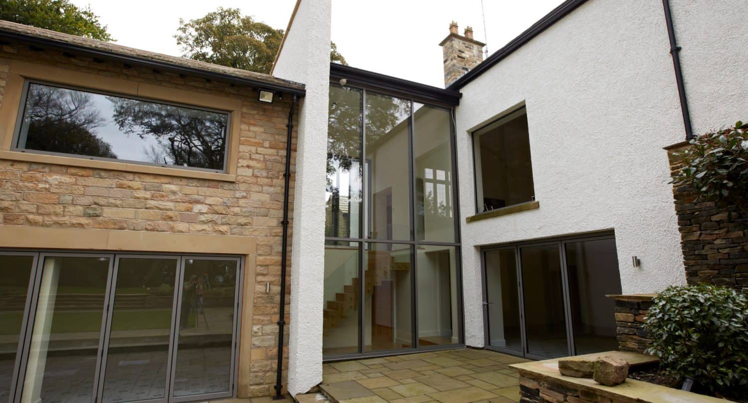 Modern house exterior with a mix of stone and white walls, large floor-to-ceiling glass windows, sliding glass doors, and a paved patio area. Trees can be seen reflected in the windows.