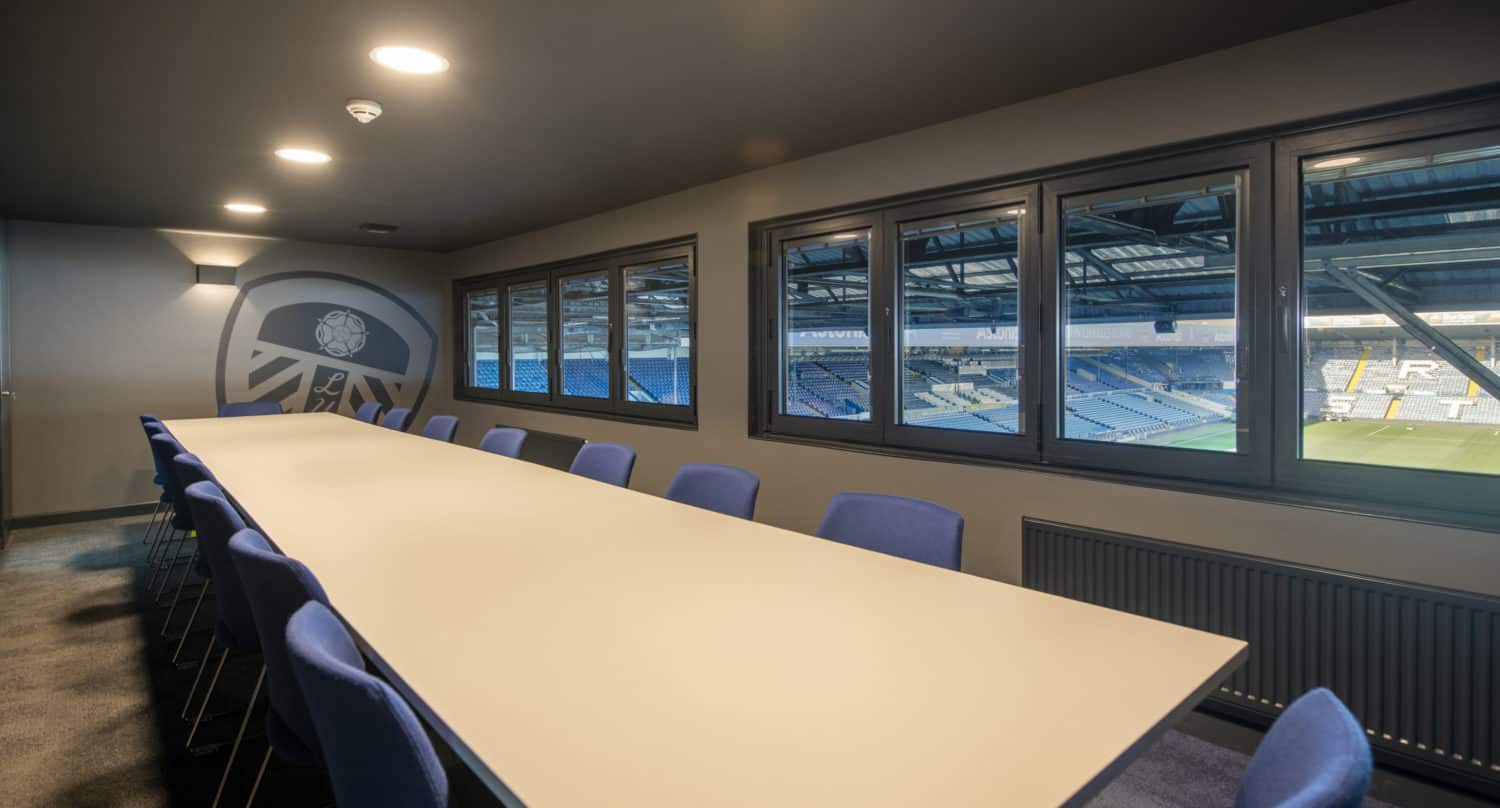 A modern conference room with a long white table, blue chairs, and large windows featuring bespoke glass solutions overlooking a stadium with rows of seats and a football field visible outside.