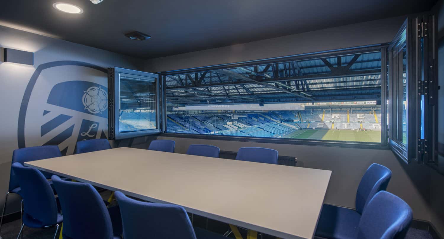 A modern meeting room with blue chairs and a long white table overlooks a large football stadium through open sliding doors, providing a clear view of the empty stands and pitch.