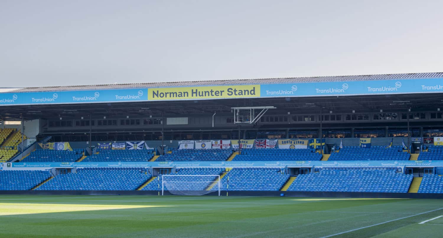 A view of empty blue seats and a goalpost at a football stadium, with a large sign reading Norman Hunter Stand above the seating area. The stand features glass roofing, while various flags are displayed along the back wall.