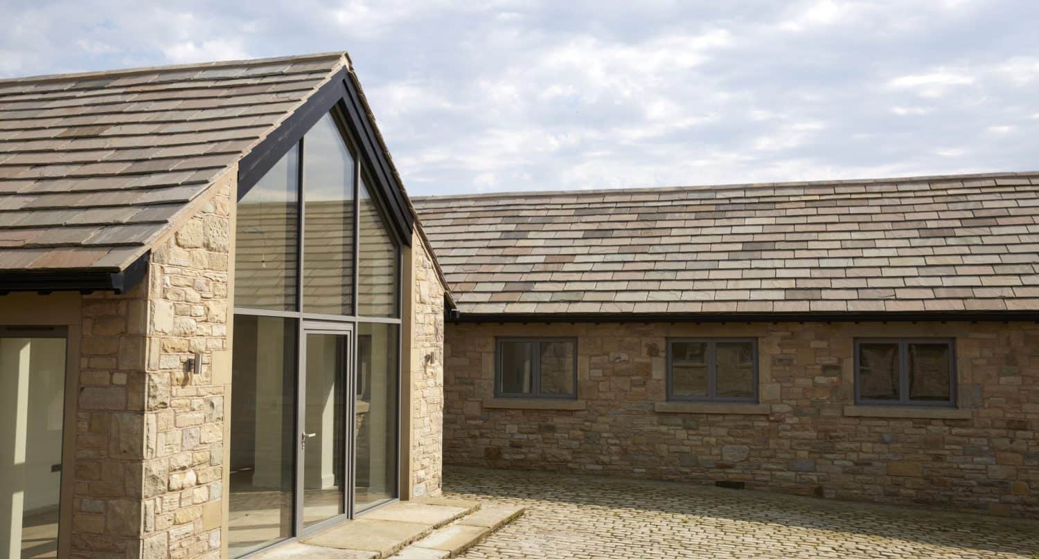 A modern stone building with bespoke glass solutions, large glass doors and windows, sloped roofs with grey tiles, and a cobblestone courtyard under a partly cloudy sky.