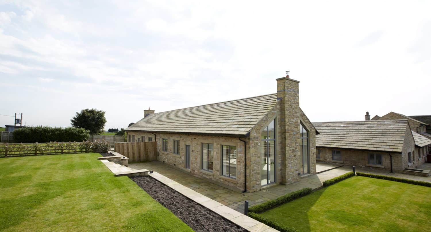 Modern stone house with large windows and sliding doors, sloped roof, neatly trimmed lawn, stone patio, and garden beds, set in a rural landscape under a bright, partly cloudy sky.