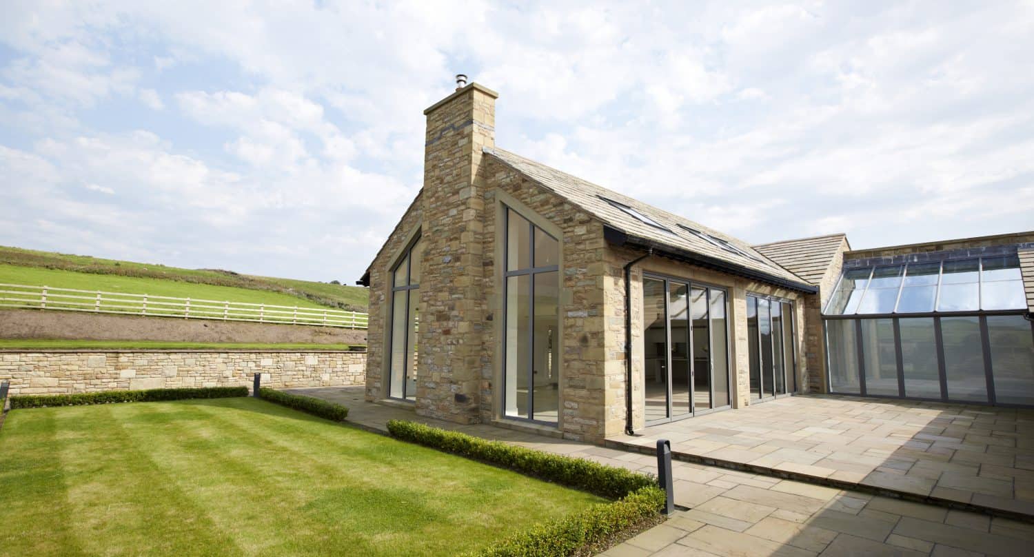 A modern stone house with bespoke glass solutions, featuring large windows and doors, a chimney, and a patio. The neatly trimmed lawn and hedges surround the home, which overlooks fields with a white fence under a partly cloudy sky.